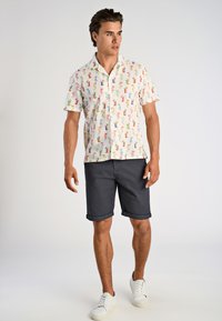 Kortærmet skjorte med farverigt cocktailmønster på en lys baggrund, kombineret med mørke shorts med rullet kant og hvide sneakers.