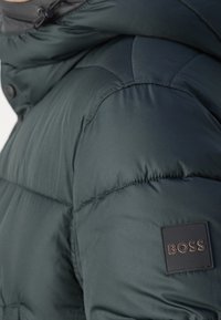 Primo piano della spalla di una giacca imbottita verde scuro con una toppa quadrata nera etichettata "BOSS" sulla parte superiore del braccio.