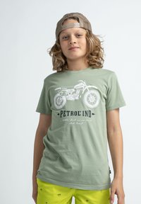 Zelené tričko s krátkým rukávem, které má bílou grafiku motocyklu a text. Materiál vypadá měkký; volný střih. Kombinujte s jasně žlutými šortkami.