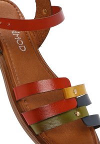 Sandalias planas coloridas con correas de cuero en múltiples capas en rojo, amarillo, verde y azul marino. Incluye una suela marrón texturizada y acentos dorados.