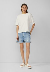 Kremowa bawełniana koszulka, luźny fason, połączona z wytartymi jasnoniebieskimi szortami dżinsowymi. Obuwie to grube białe klapki na platformie.