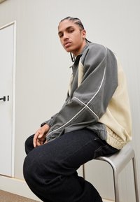 Lys grå og beige lynlåsejakke med elastiske manchetter og kant; kombineret med mørke denimjeans. Glat tekstur og struktureret design.
