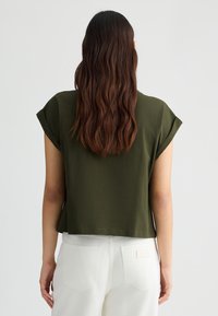 Top corto verde scuro a maniche corte con vestibilità rilassata, tessuto testurizzato e orlo dritto. Indossato con pantaloni bianchi a vita alta.