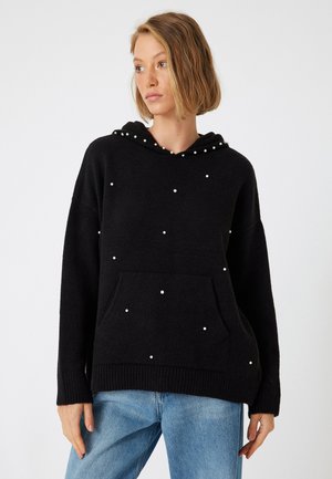 Zwarte gebreide hoodie met een voorzak, versierd met kleine witte parels, en geribbelde zoom en boorden. Draag hem over lichtblauwe jeans.
