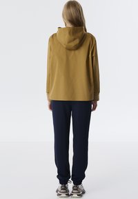 Waterdichte jas in mosterdkleur met een capuchon; gecombineerd met marineblauwe joggers, elastische manchetten en grijs-witte sneakers. Strak ontwerp en pasvorm.