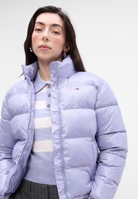 Chaqueta acolchada de color púrpura claro con cuello alto, que cuenta con cierre de cremallera en el frente y detalle del logo en el pecho. Textura suave y brillante con capas claras en contraste.