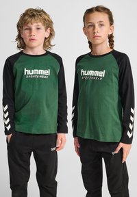 Magliette di cotone verde a maniche lunghe con maniche nere, con logo bianco e motivi a chevron. Abbinate a pantaloni neri, anch'essi con logo.