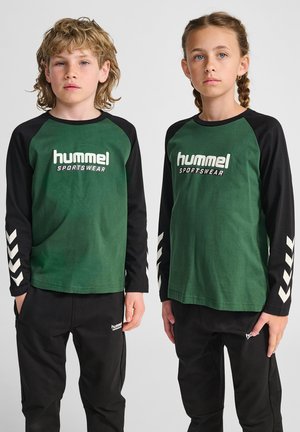 Magliette di cotone verde a maniche lunghe con maniche nere, con logo bianco e motivi a chevron. Abbinate a pantaloni neri, anch'essi con logo.