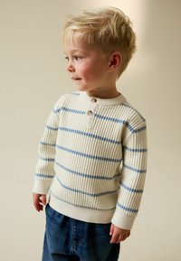 Maglione color crema con lavorazione a maglia testurizzata, caratterizzato da strisce orizzontali blu e scollo henley con bottoni. Abbinato a jeans denim blu.