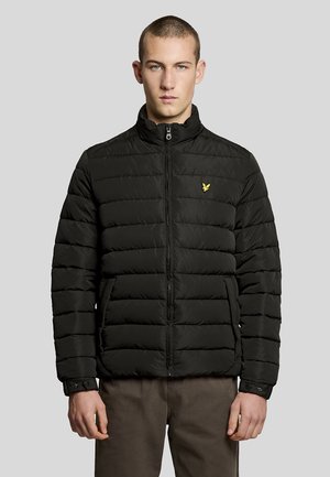 Joven con una chaqueta acolchada negra con un logo de pájaro amarillo en el pecho, de pie frente a un fondo liso de color gris claro.