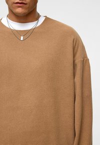 Bruine gestructureerde pullover met een losse pasvorm, voorzien van een ronde hals. Gelaagd over een wit shirt, aangevuld met gelaagde zilveren kettingen.