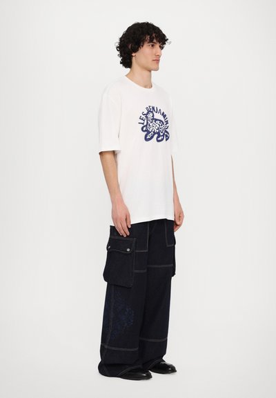 Λευκό βαμβακερό T-shirt με μπλε γραφικό σχέδιο, συνδυασμένο με σκούρες τζην cargo παντελόνες με πολλαπλές τσέπες και διακριτές σχέδιο λεπτομέρειες.