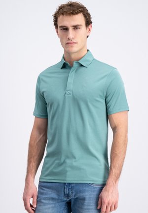 Man met een turquoise polo met korte mouwen en blauwe spijkerbroek, staand tegen een effen lichte achtergrond, kijkend naar de camera.