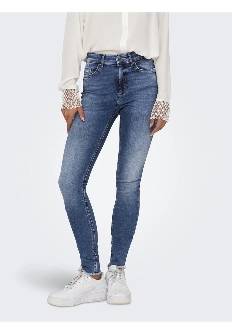 Jean skinny bleu avec un effet délavé, cinq poches et ourlet effiloché, associé à un chemisier blanc à manches longues avec des accents en dentelle. Baskets blanches.