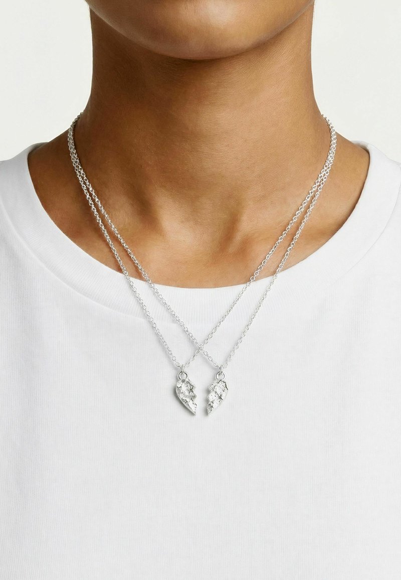 Deux colliers en argent avec des pendentifs assortis en forme de cœur brisé portés par une personne à la peau mate et portant une chemise blanche.