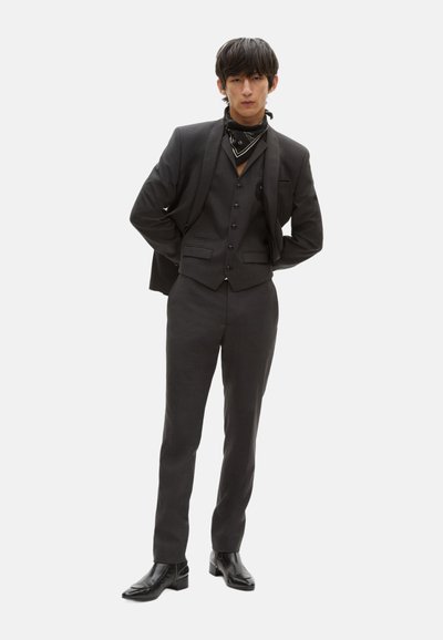 Maßgeschneiderter Dreiteiler aus dunkelgrauem Stoff, bestehend aus einer Weste mit Knöpfen, enganliegenden Hosen und schwarzen Leder-Ankle-Boots.