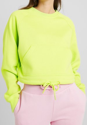 Person iført en neongul cropped sweatshirt med forlomme og pink sweatpants med snøre, hænder i lommerne.