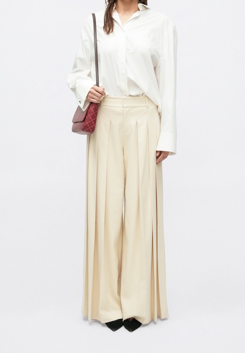 Femme portant une chemise blanche ample et un large pantalon plissé beige, tenant un petit sac à main rouge, chaussures noires pointues visibles.
