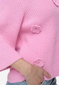 Světle růžový pletený svetr s žebrovanou texturou, zdobený dekorativními květinovými aplikacemi a širokými rukávy, doplněný džínovými kalhotami.
