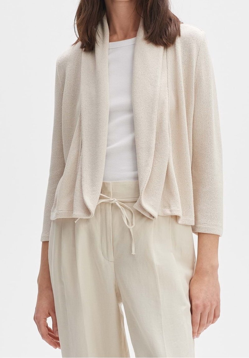 Femme portant un cardigan beige clair ouvert sur le devant, un haut blanc côtelé et un pantalon ample crème à cordon, vue du cou aux hanches.