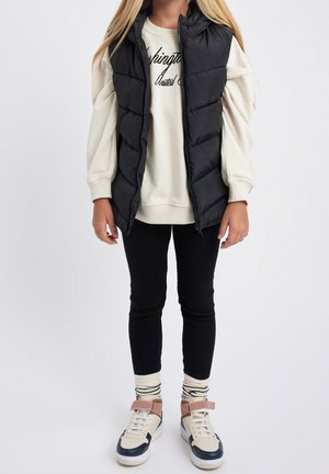 Enfant portant un gilet matelassé noir, un sweat crème avec un texte, des leggings noirs, des chaussettes rayées et des baskets blanches avec des bandes roses sur fond uni.