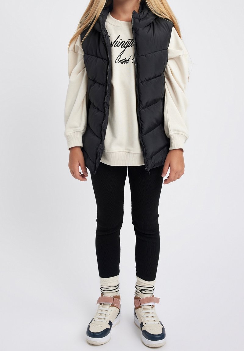 Enfant portant un gilet matelassé noir, un sweat crème avec un texte, des leggings noirs, des chaussettes rayées et des baskets blanches avec des bandes roses sur fond uni.