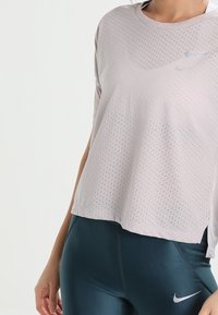 Camiseta deportiva de color gris claro, perforada, con mangas cortas y corte holgado. Presenta un pequeño logo reflectante de Nike en el frente.