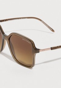 Sunčane naočale od acetata s smeđim gradientnim lećama, kvadratnog oblika i prozirnim smeđim okvirom. Imaju metalne detalje s logotipom "A/X" na stranama.