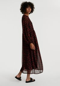 Schwarzes, durchsichtiges Maxikleid mit rotem Blumendruck, langen Ärmeln und einem lockeren Schnitt. Getragen mit schwarzen Riemensandalen.