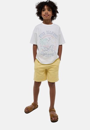 T-shirt en coton blanc avec un motif coloré inspiré de la plage, associé à un short jaune clair et des sandales brunes avec une bride ajustable.