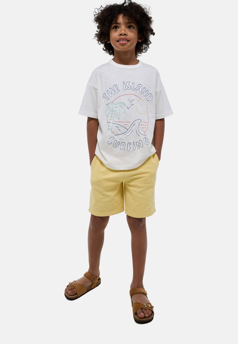 T-shirt en coton blanc avec un motif coloré inspiré de la plage, associé à un short jaune clair et des sandales brunes avec une bride ajustable.