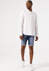 Camisa blanca de lino de manga larga, pantalones cortos de mezclilla azules con dobladillos enrollados y zapatillas blancas. Tatuajes notables en las piernas, diseño casual y sencillo.