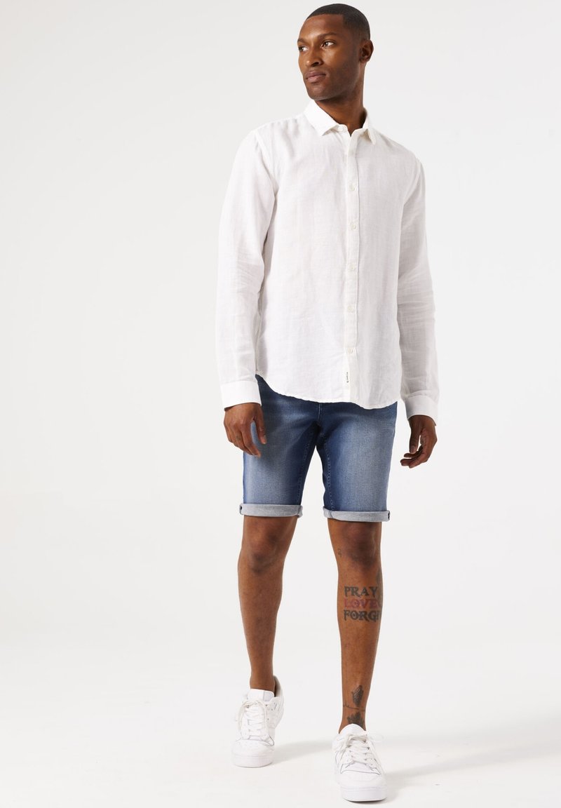 Camisa blanca de lino de manga larga, pantalones cortos de mezclilla azules con dobladillos enrollados y zapatillas blancas. Tatuajes notables en las piernas, diseño casual y sencillo.