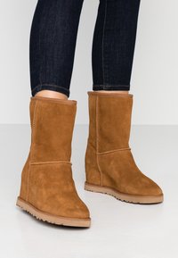 Botines de ante marrón con cuña, llevados con jeans skinny oscuros sobre un suelo blanco y fondo sencillo.