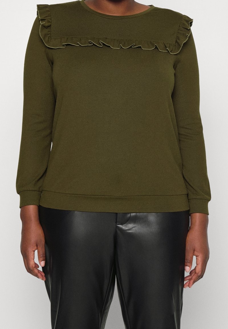 Haut vert olive foncé à manches longues avec un empiècement volanté, associé à un pantalon en similicuir noir, porté par une personne sur un fond uni.