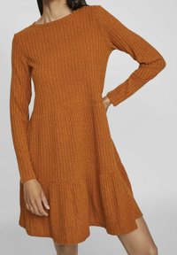 Femme portant une robe en maille côtelée orange brûlé à manches longues avec une jupe évasée, sur un fond gris clair uni.