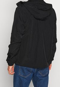 Mies, joka pukeutunut mustaan hupulliseen takkiin ja sinisiin farkkuihin, kuvattuna takaa yksinkertaisen vaalean taustan edessä.