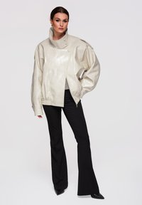 Donna che indossa una giacca oversize in pelle beige lucida con collo alto, abbinata a pantaloni neri svasati e scarpe nere con punta affusolata, in piedi con la mano in tasca.