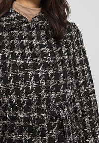 Svart och vitt mönstrad kappa gjord av texturerad tyg, med dubbelknäppning, stora svarta knappar och midjebälte.