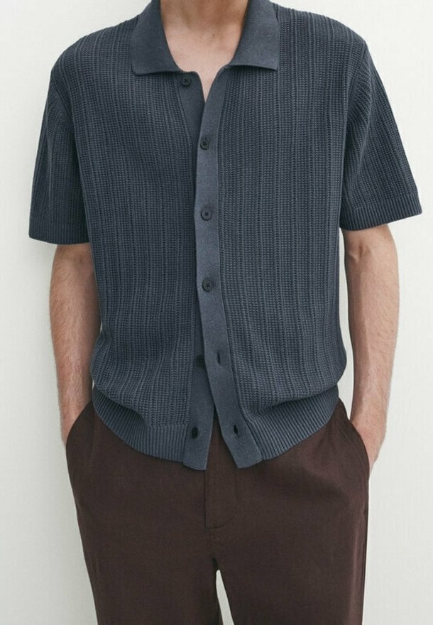 Homme portant une chemise à boutons à manches courtes en tricot gris foncé avec col et un pantalon marron foncé, les mains dans les poches.