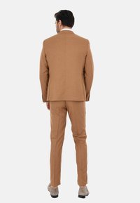 Completo marrone su misura con una texture liscia, rever a scollo e pantaloni dritti, abbinato a scarpe grigie chiare.