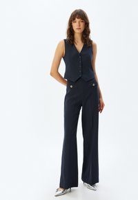 Marinblå randig väst med V-hals och knappar, tillsammans med högmidjade vida byxor med metalliska accenter. Silverfärgade skor fulländar looken.