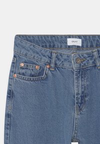 Denimbukser i lyseblå med kobbernitter, standard talje og en lille frontlomme. Har en label med teksten "GRUNT" ved taljen.