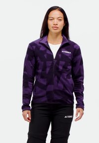 Chaqueta de forro polar morada con un patrón geométrico, cierre frontal con cremallera y una marca en el lado izquierdo del pecho, combinada con pantalones negros que presentan un logo.