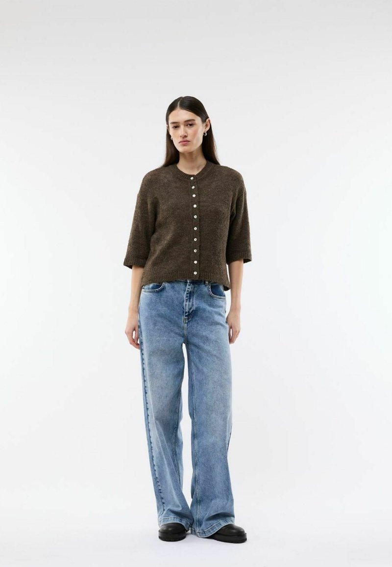 Femme aux longs cheveux foncés portant un cardigan marron boutonné, un jean bleu clair à jambes larges et des chaussures noires, debout devant un fond blanc.