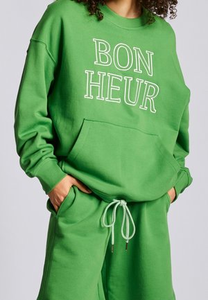 Personne portant un sweat-shirt vert vif et un short assorti avec un cordon blanc, le sweat-shirt affiche "BON HEUR" en grandes lettres blanches.