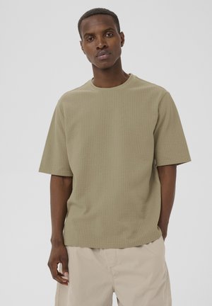 Männliches Model trägt beige texturiertes Kurzarmhemd und hellbraune Shorts, steht vor einem einfarbigen hellgrauen Hintergrund.