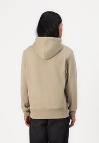 Sudadera con capucha en color beige, confeccionada en un tejido suave. Presenta un corte relajado, puños y dobladillo de canalé, y un bolsillo tipo canguro.