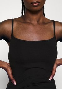 Svart linne med tunna axelremmar, fitted design, slät textur och fyrkantig halsringning. Hudton och händer som är placerade på höfterna är synliga.