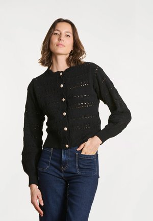 Femme aux cheveux mi-longs portant un cardigan en maille noir boutonné et un jean bleu foncé, posant avec une main dans la poche.